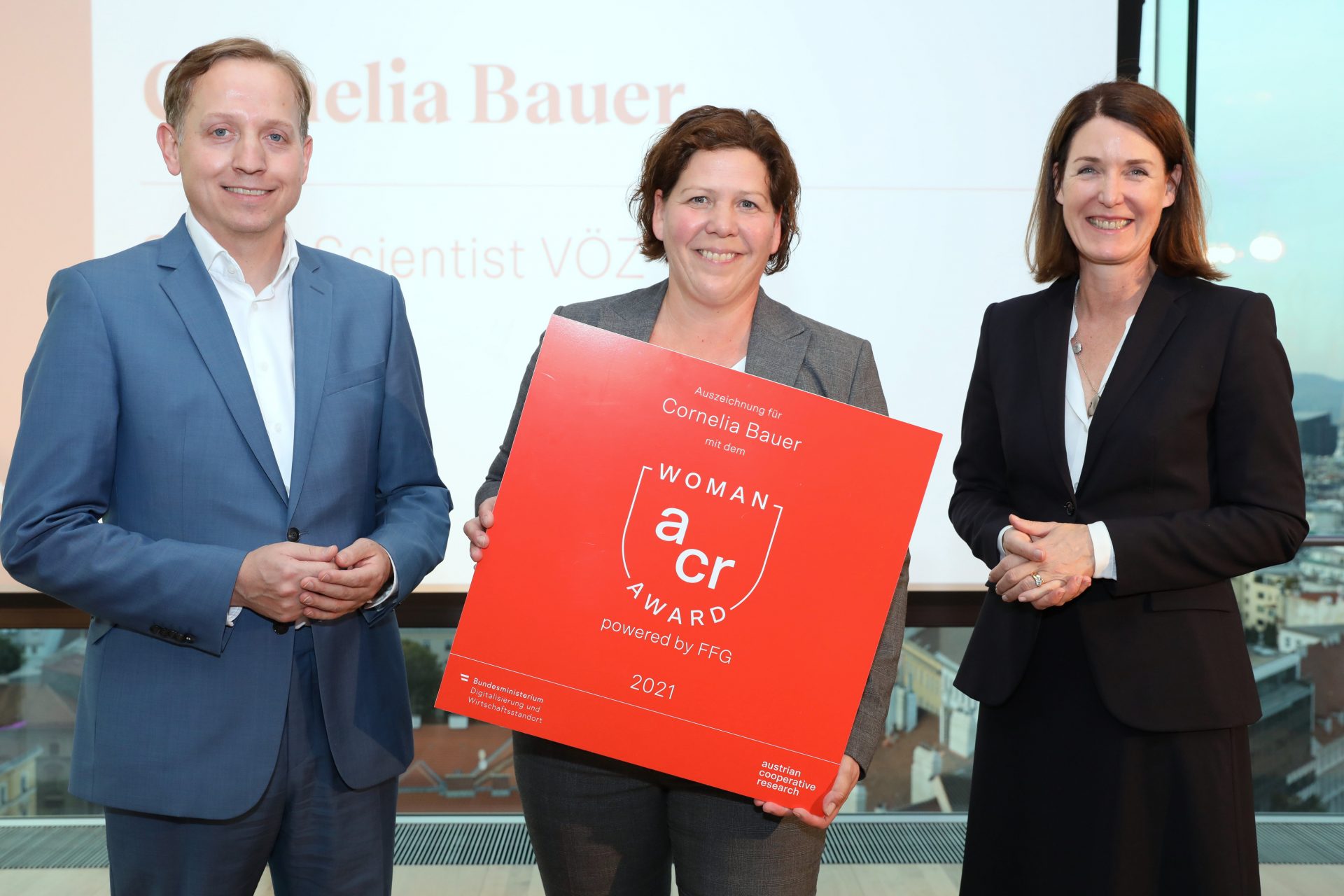 Drei Personen auf der Bühne, mittlere hält ein großes rotes Schild in der Hand