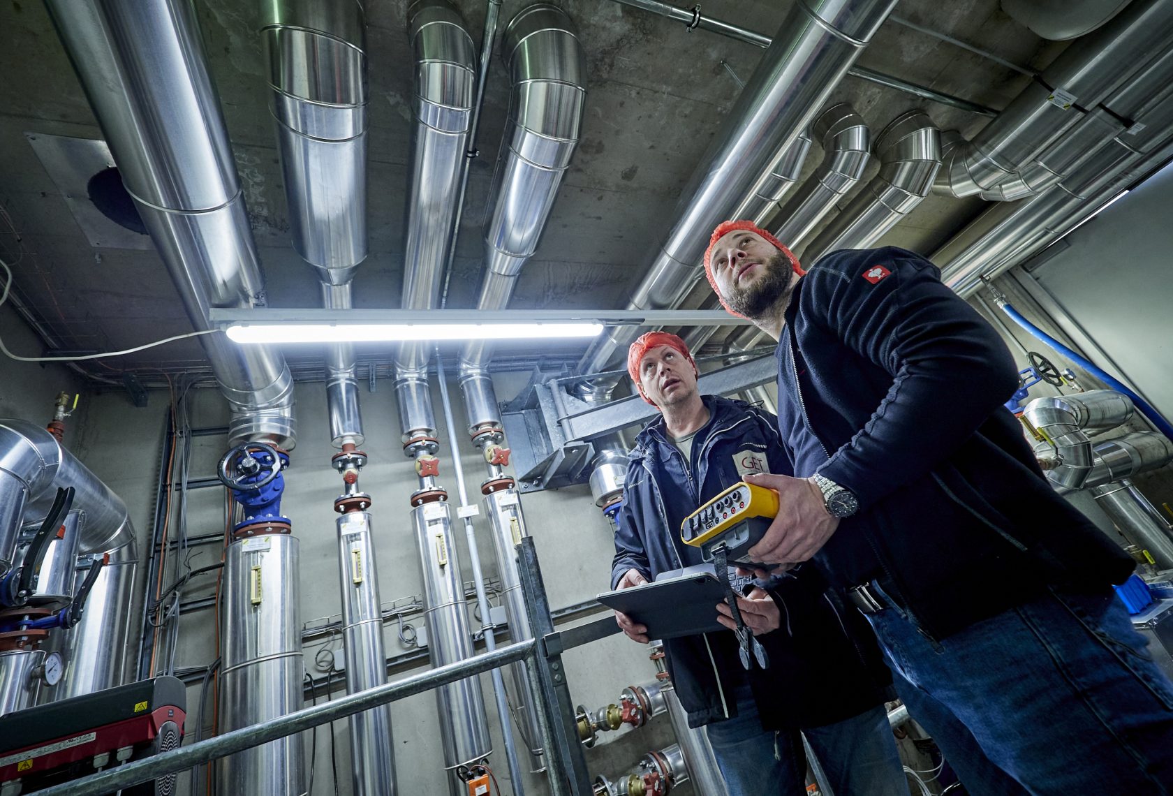 Zwei Personen mit Messgeräten vor mehreren Rohren, die an der Wand verlaufen