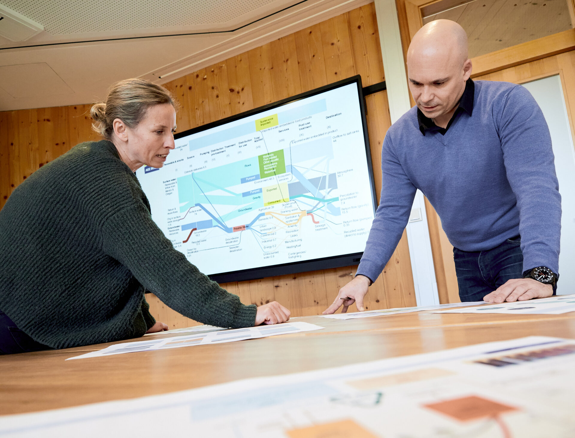 2 Personen im Gespräch, über einen Plan gebeugt und mit Leinwand im Hintergrund
