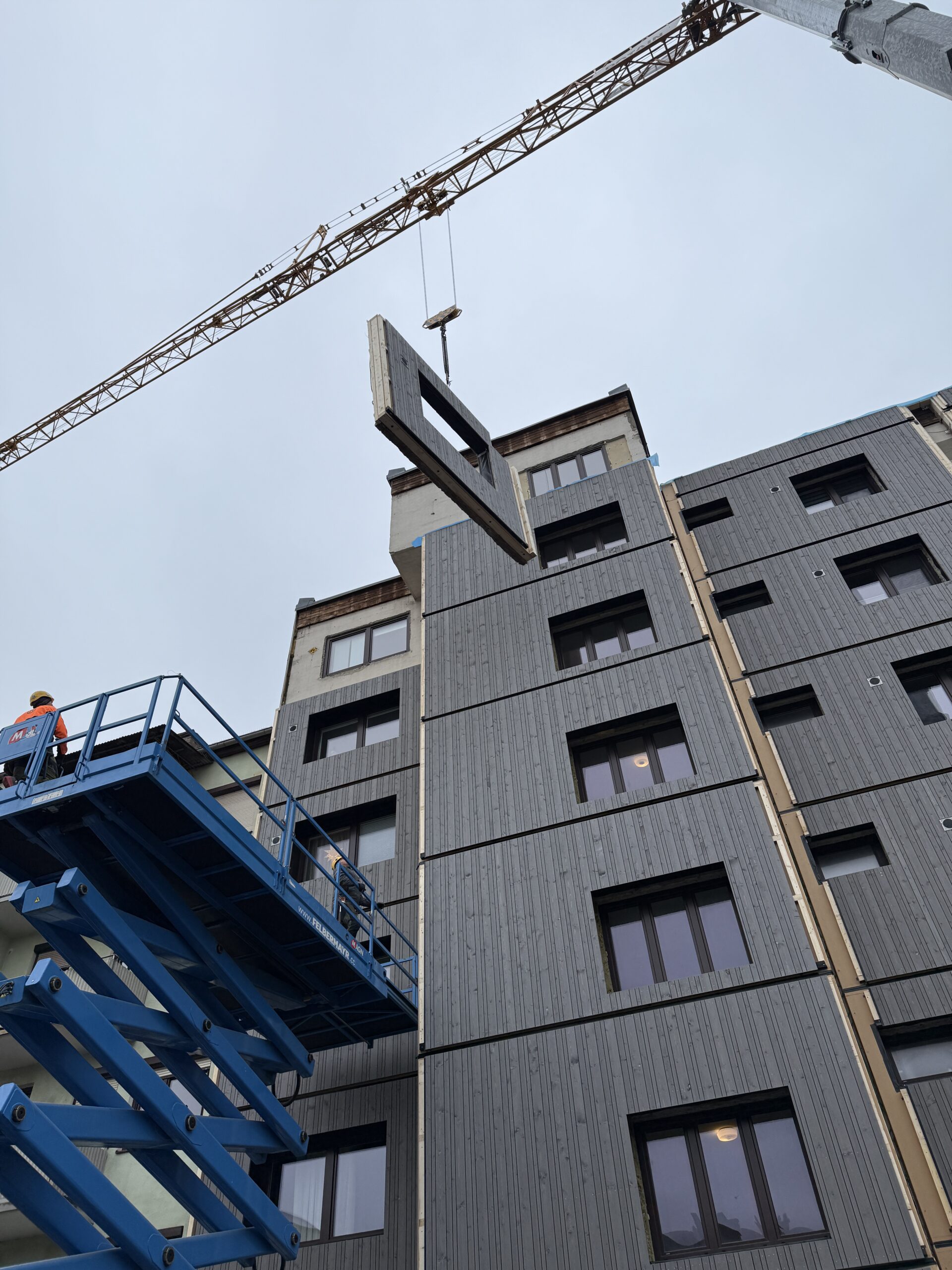 Gebäude im Hintergrund, Baukran mit Fassadenelement im Vordergrund